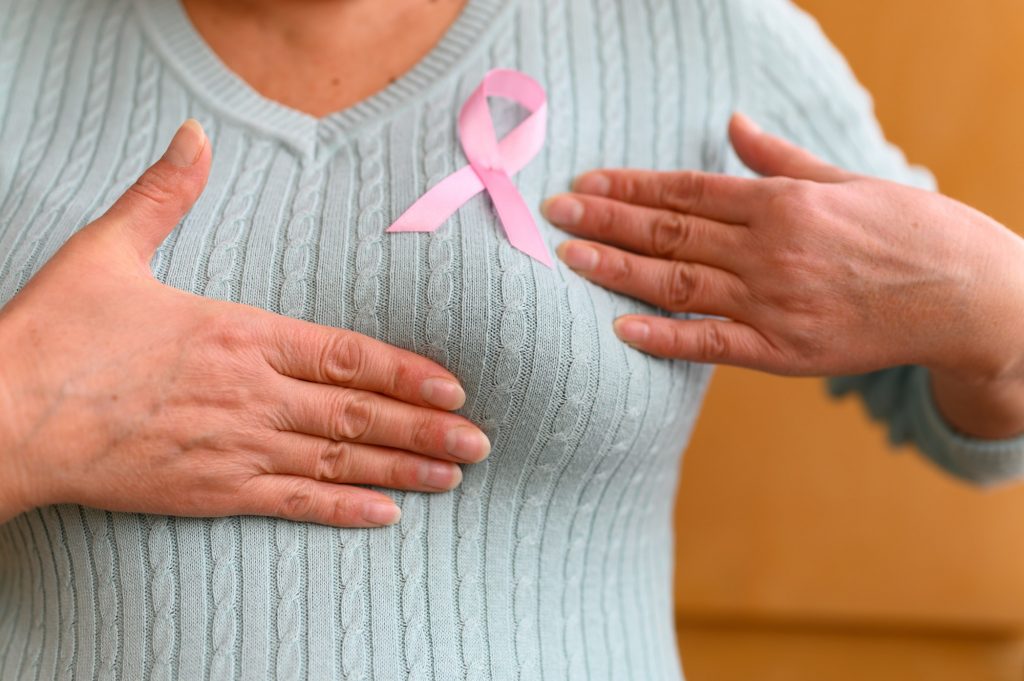 hands of a mature woman examine her breasts. a pink badge ribbon is attached to a knitted sweater